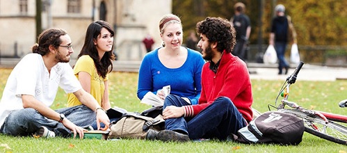 A group of 4 students sitting on grass, some have open books, one has a bike.